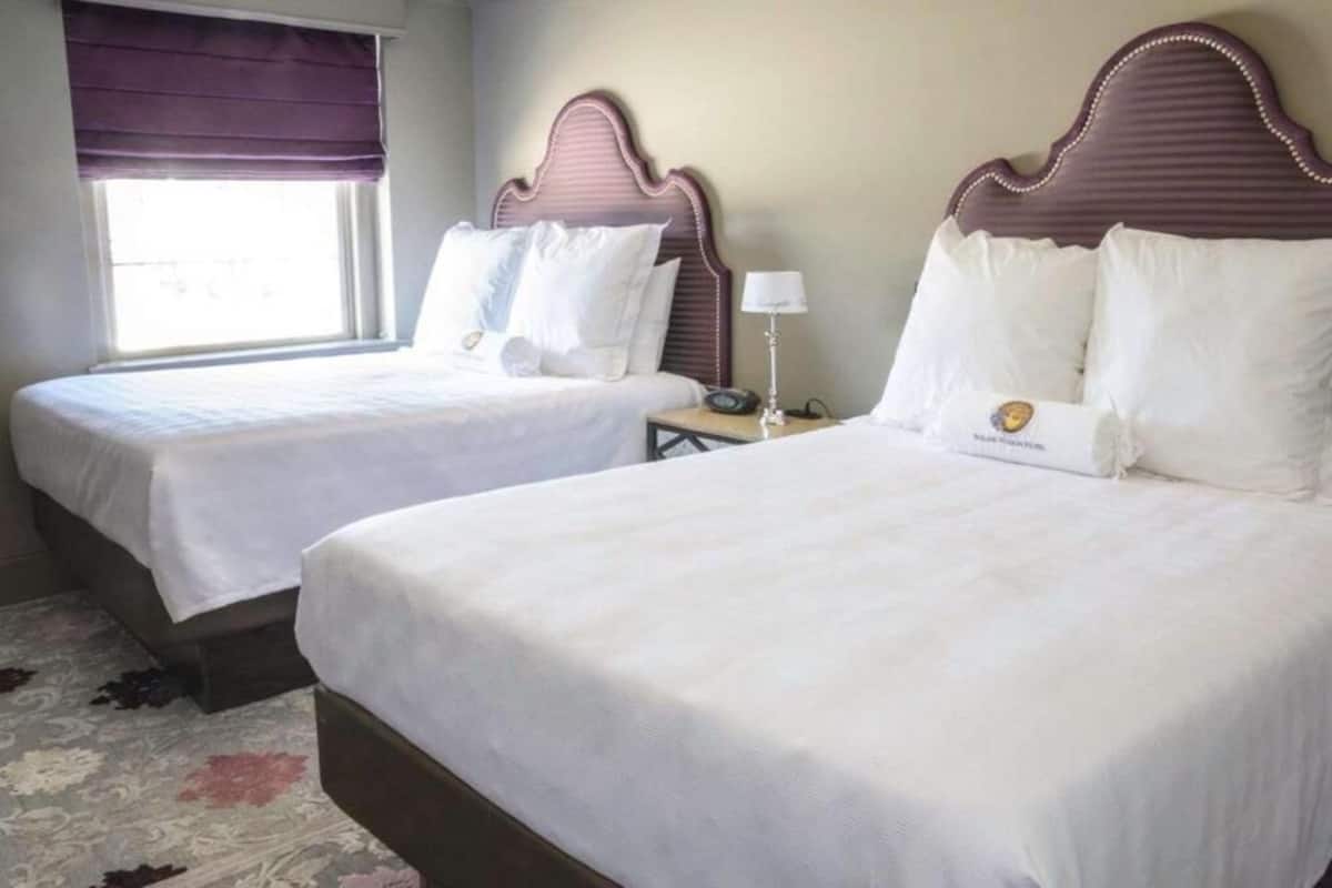 A hotel room with two beds, each with white linens and headboards. There is a window with a purple blind, a bedside table with a lamp, and a small clock.