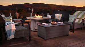 Une terrasse dotée de meubles en osier, d’une table ronde agrémentée d’un foyer, et offrant une vue panoramique sur des collines en arrière-plan.