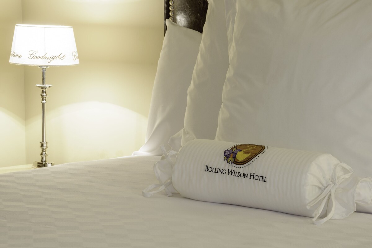 Una cama cuidadosamente hecha con una almohada que dice "Bolling Wilson Hotel" y una lámpara con la palabra "Buenas noches" en su pantalla.