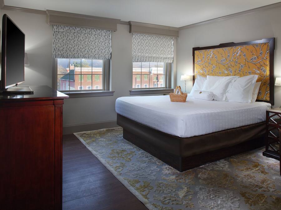 A hotel room with a large bed, a wooden dresser, a television, and two windows with patterned curtains.