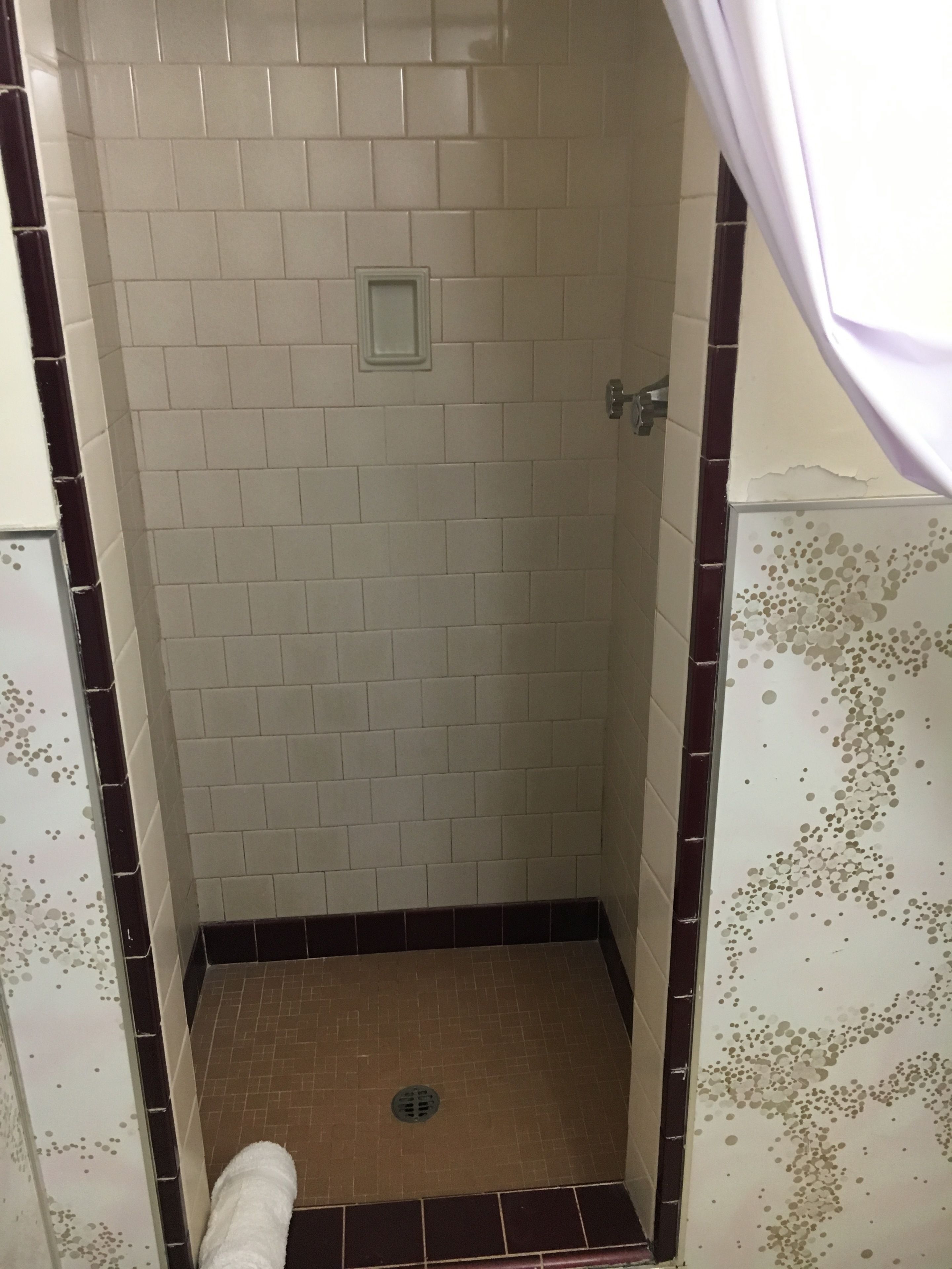 A small, enclosed shower area with white and brown tiles, a drain, and a door with a metal handle.