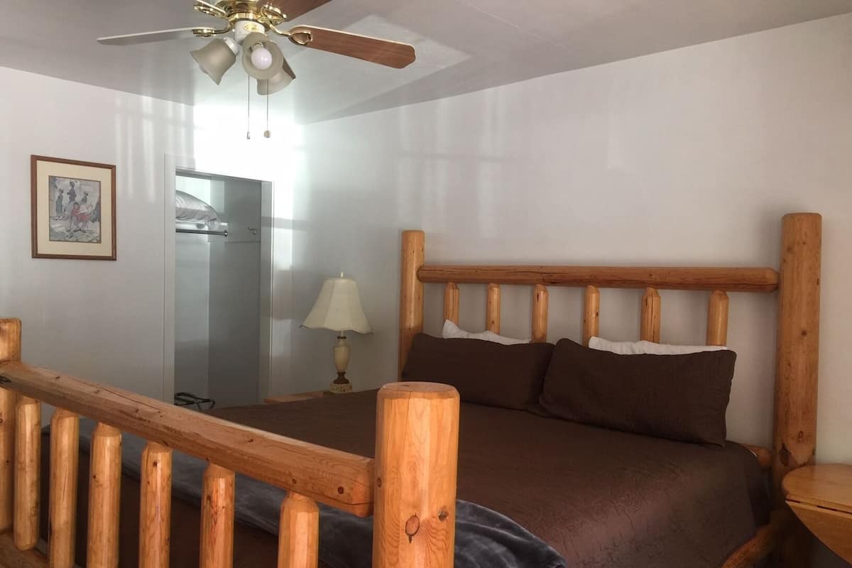 A bedroom with a wooden bed frame, a ceiling fan, a bedside lamp, and a framed picture on the wall.
