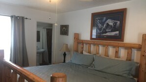 Un dormitorio con cabecera de madera, cama con cubrecama gris, mesita de noche, lámpara, un cuadro enmarcado en la pared, y un baño visible a través de una puerta abierta.