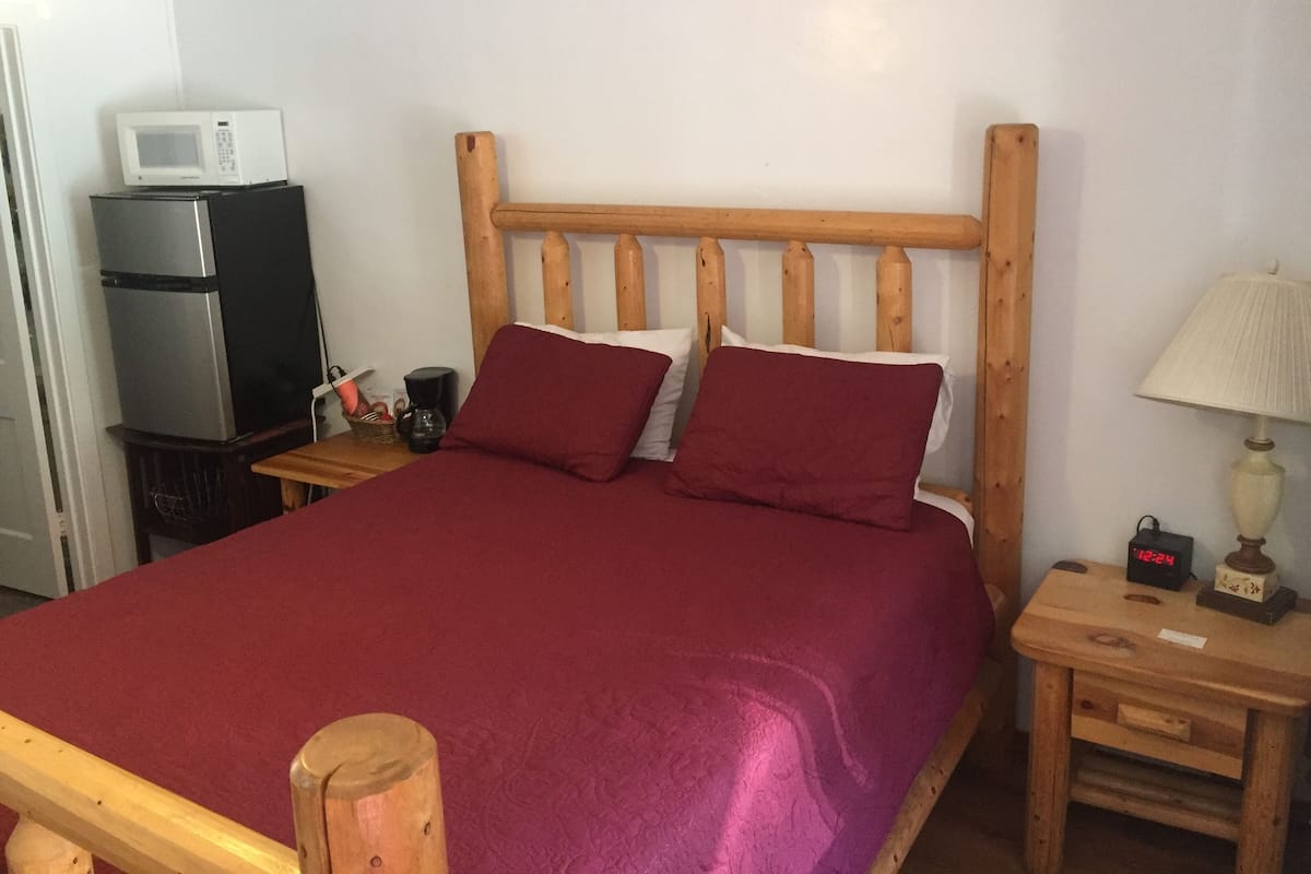 A bed with a wooden headboard, a red bedspread, two pillows, a bedside table with a lamp, a clock, and a microwave on a refrigerator.