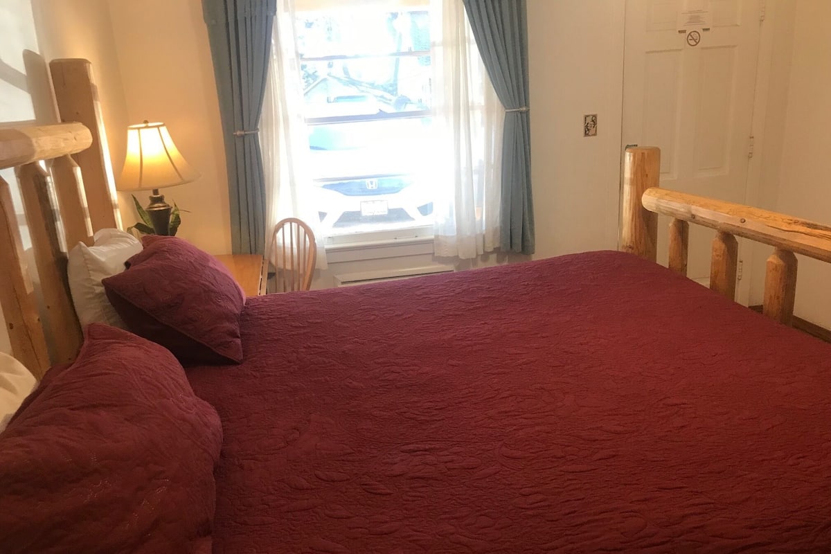 A bedroom with a large bed, a wooden headboard, a nightstand with a lamp, and a window with curtains.