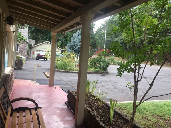 A covered porch with a bench, a parking lot, and a view of a residential street with houses and greenery.