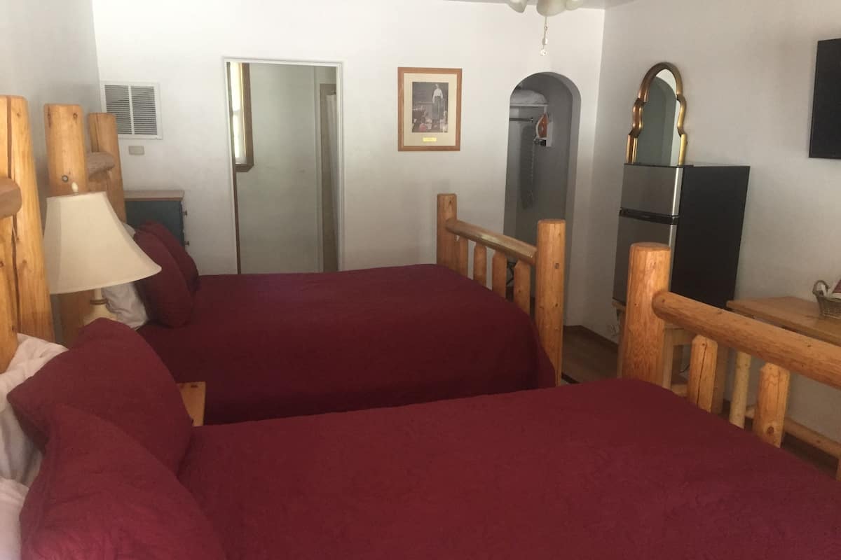 A room with two beds, a wooden headboard, a desk with a mirror, a framed picture on the wall, and a door leading to another room.