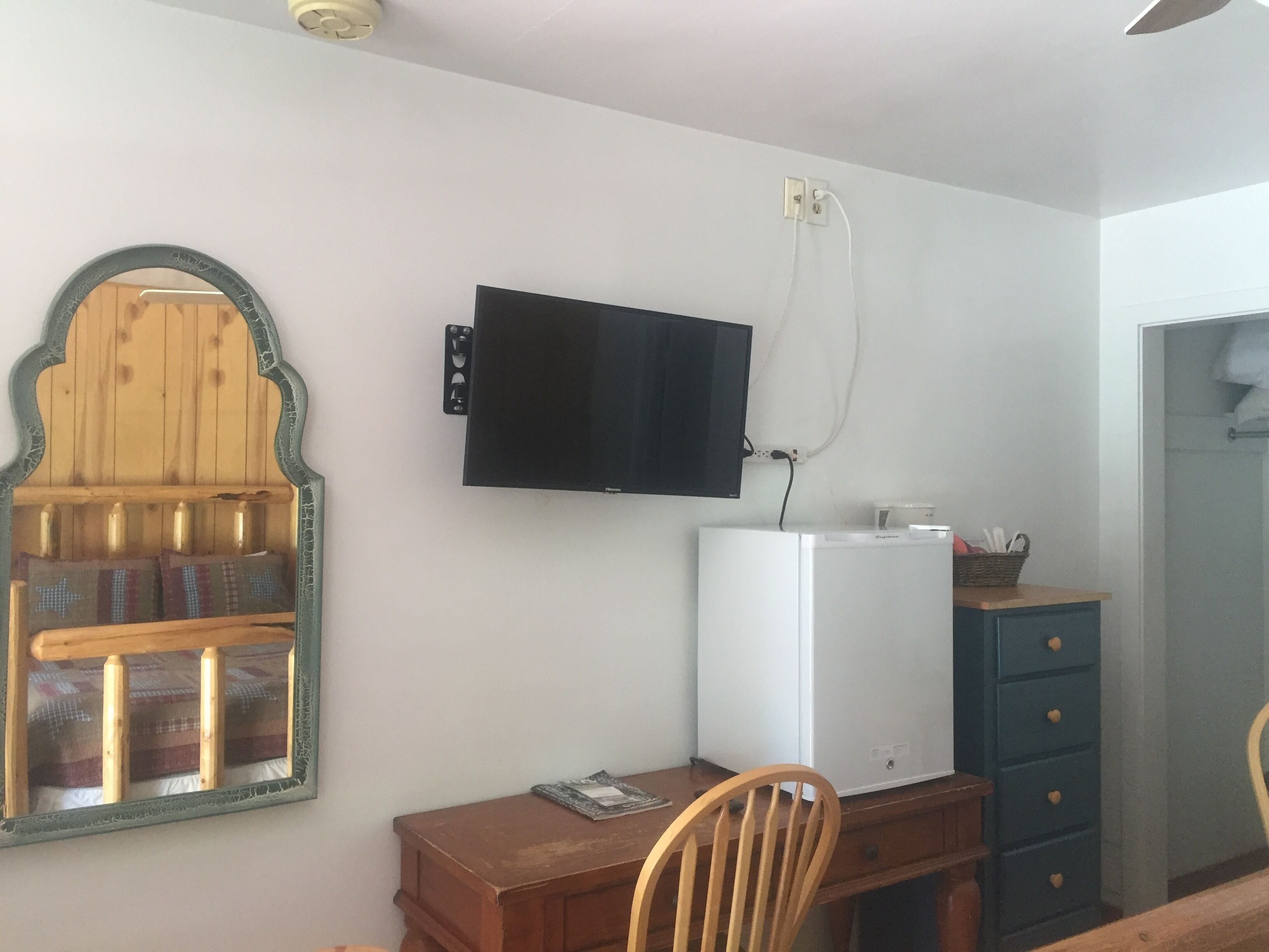 Une pièce comprenant un bureau en bois, une chaise, un téléviseur fixé au mur, un réfrigérateur blanc et un meuble bleu.