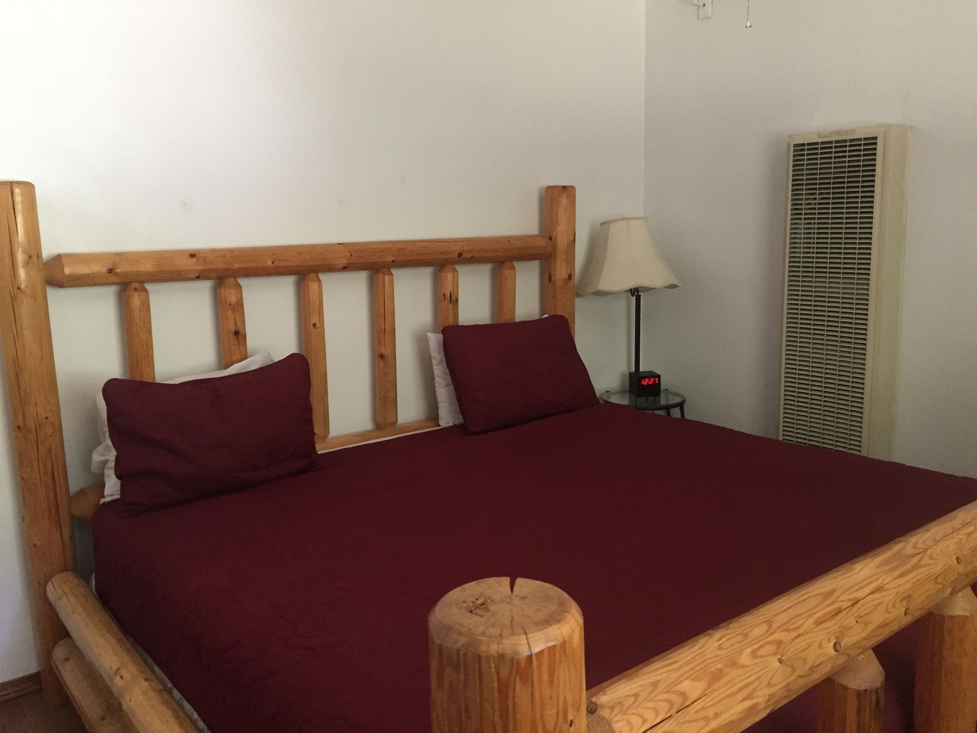 A wooden bed with a headboard, a red bedspread, and a nightstand with a lamp and a clock.