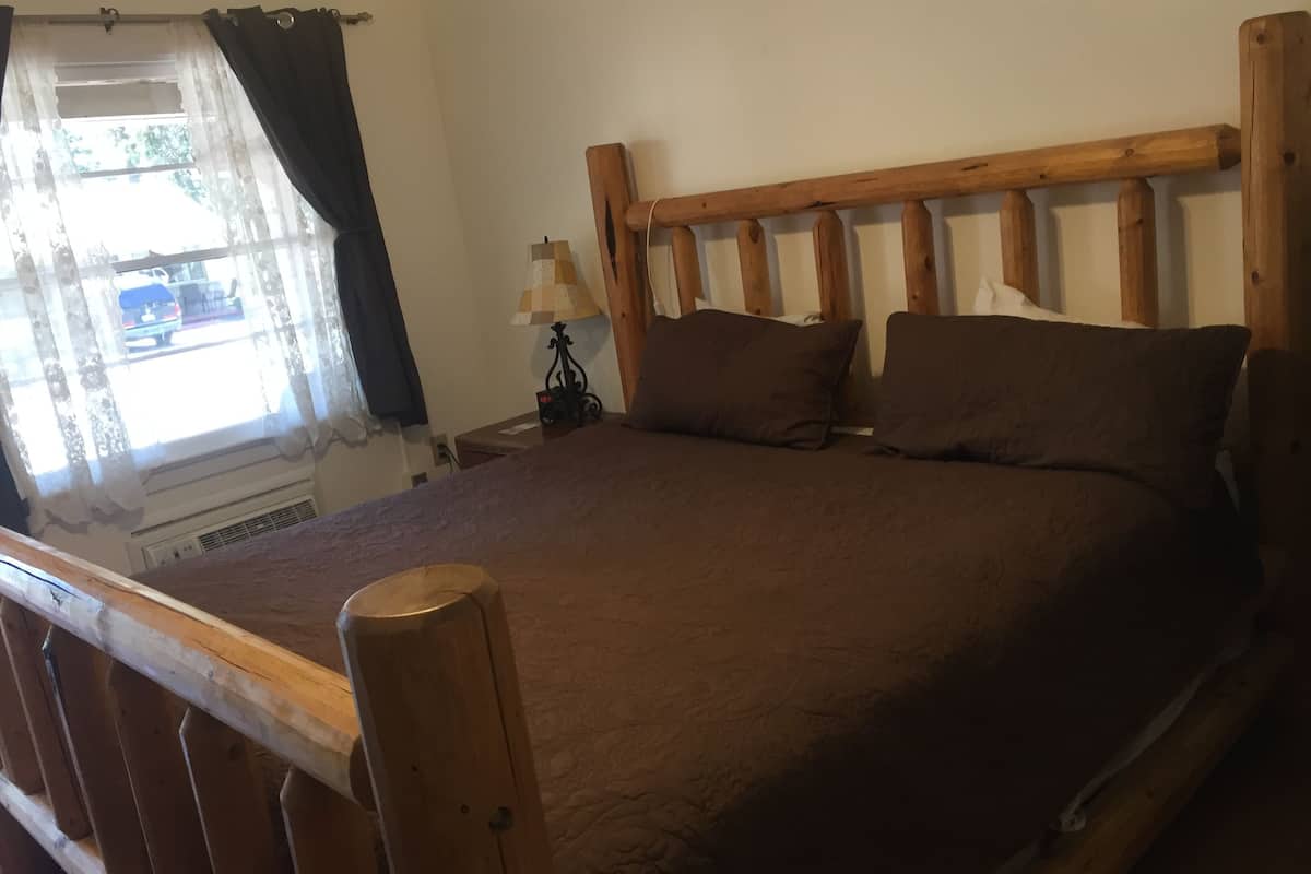 A bedroom with a wooden headboard, a bed with brown bedding, a nightstand with a lamp, and a window with curtains.