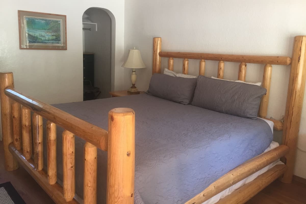 A wooden-framed bed with a grey bedspread and pillows, a wooden headboard, a bedside lamp, and a framed picture on the wall.