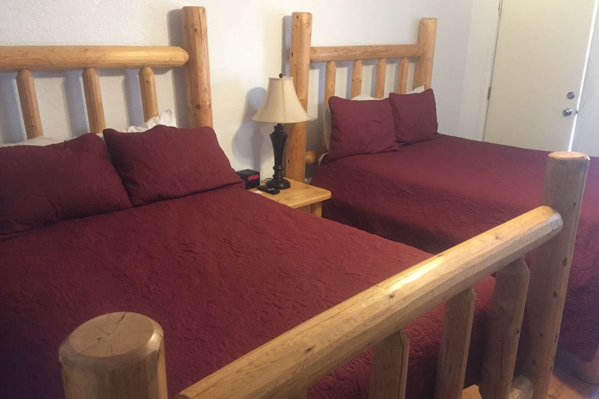 A room with two wooden beds, each with a red bedspread and matching pillows. There is a wooden headboard, a small wooden nightstand with a lamp, and a door in the background.