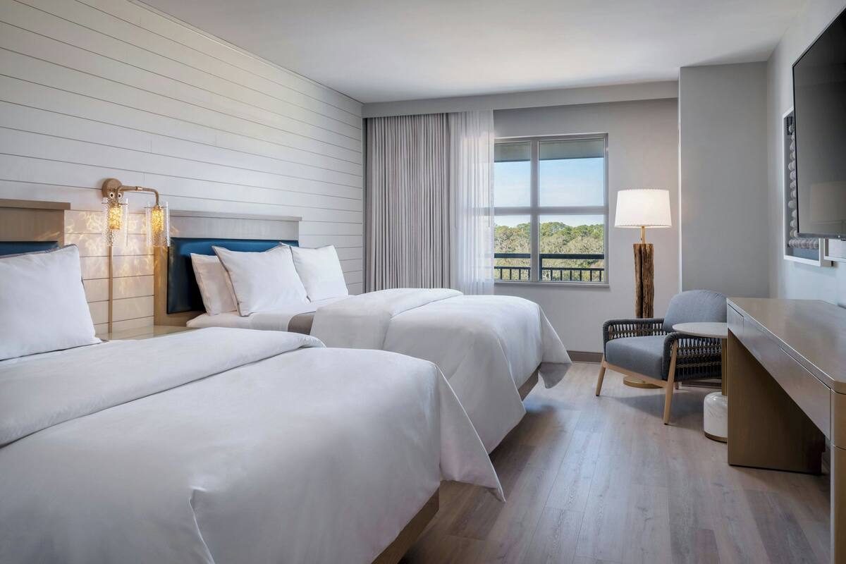 Une chambre d’hôtel avec deux lits, un bureau, une chaise, une fenêtre donnant sur un beau paysage et une lampe.