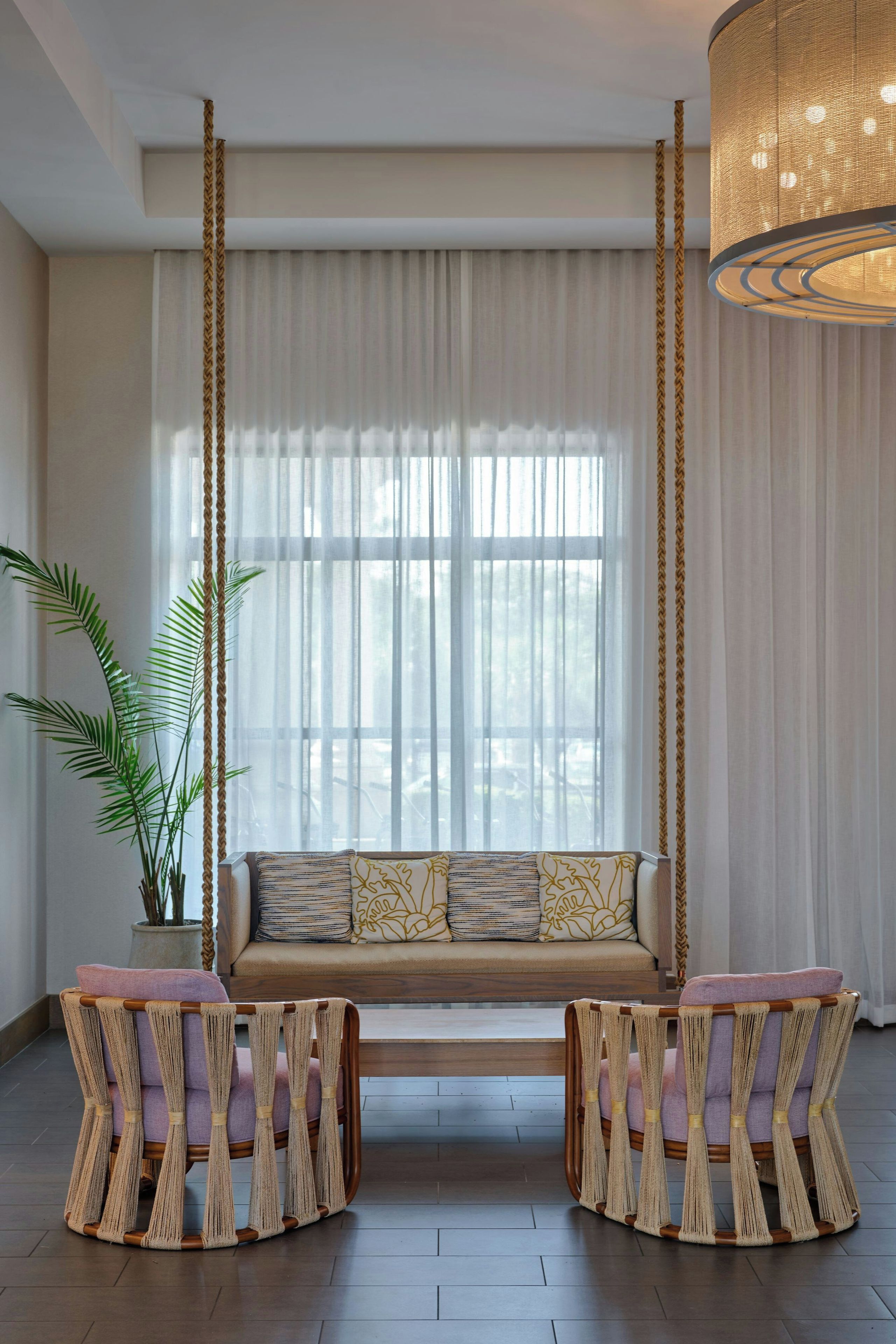 A modern living room with a sofa, two armchairs, a hanging light fixture, and a large window with sheer curtains.