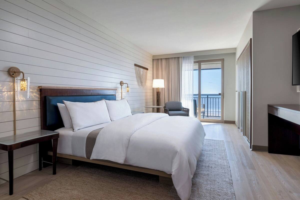 Une chambre d’hôtel avec un grand lit, un bureau, une chaise, une télévision et un balcon avec des rideaux.