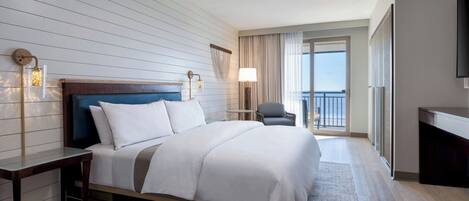 A hotel room with a large bed, a desk, a chair, a TV, and a balcony with curtains.