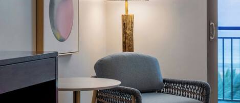 A modern living room with a grey sofa, a round table, and a lamp. There are abstract paintings on the wall and a view of the outdoors through a balcony door.