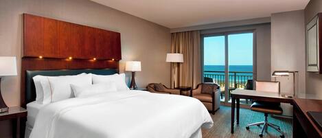 A hotel room with a large bed, a desk, a chair, a lamp, and a view of the ocean.