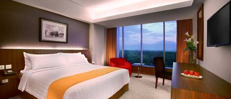 A hotel room with a large bed, a desk with a vase of flowers, a chair, a TV, and a view of the cityscape through large windows.