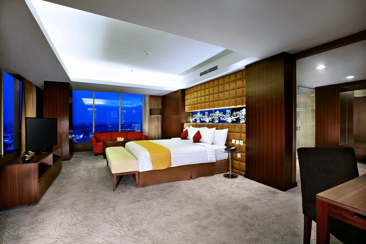 A modern hotel room with a large bed, a TV, a seating area, and a view of the city at night.