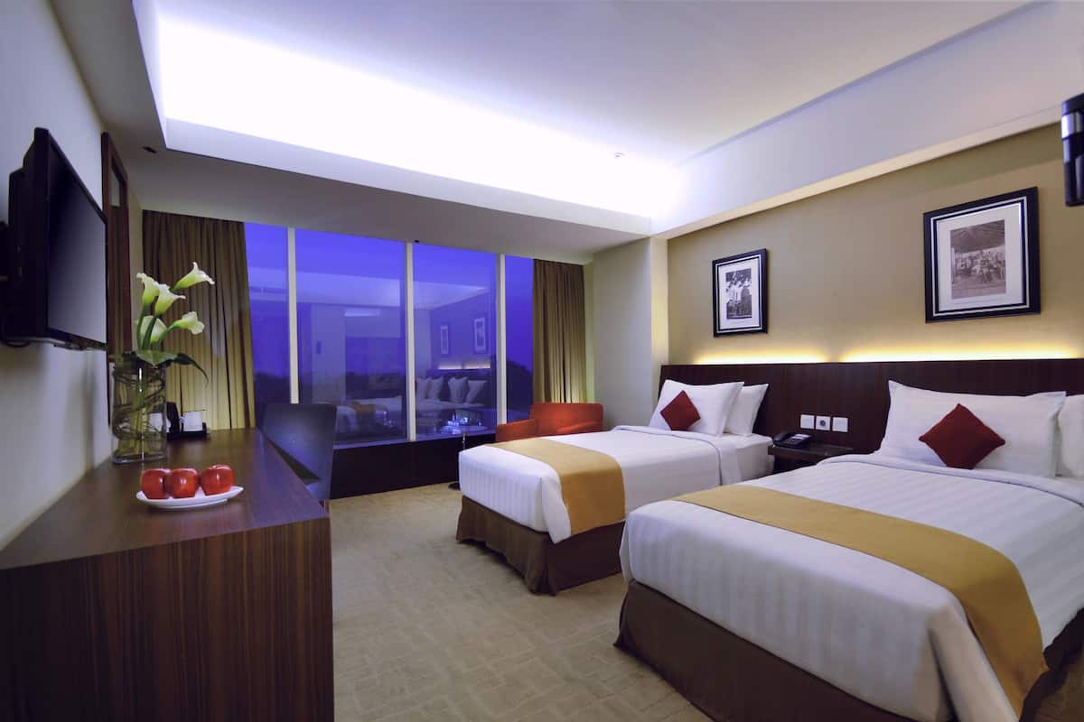 A hotel room with two beds, a desk with a plate of tomatoes, a TV, and a large window with curtains.