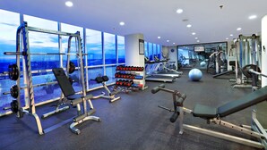 A gym with various exercise equipment, large windows, and a view of the cityscape. - Aston Madiun Hotel & Conference Center (Madiun)