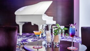 Un área de bar con un piano al fondo, tres bebidas sobre una mesa y un letrero de neón.