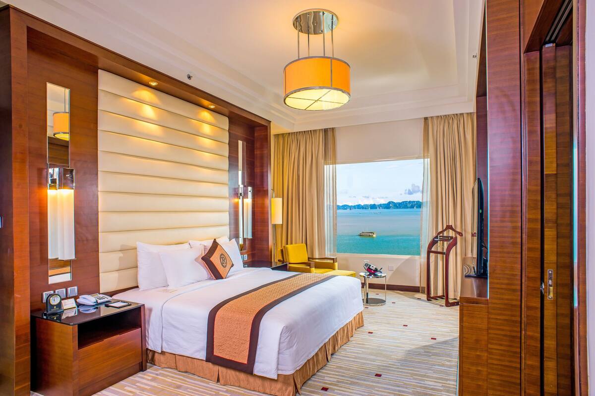 A hotel room with a large bed, a bedside table, a chair, a desk, and a view of the sea.