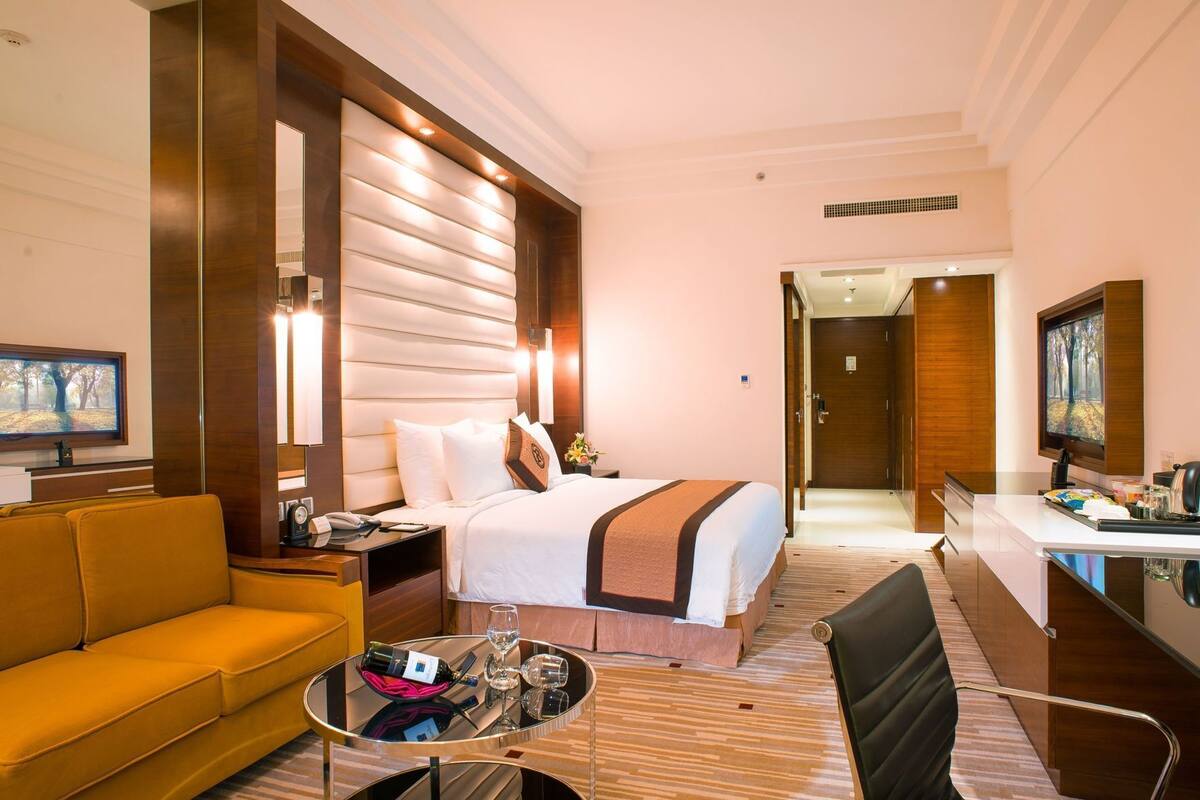 A hotel room with a bed, a sofa, a desk, a TV, and a painting on the wall.