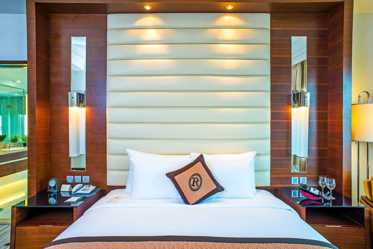 A hotel room with a large bed, a nightstand with a clock, a mirror, and a bedside table with a lamp and glasses.