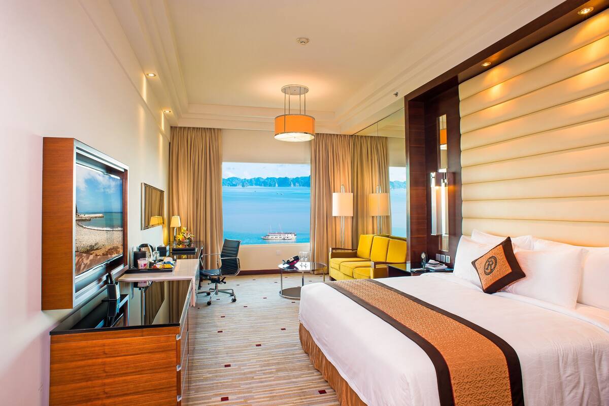 A hotel room with a bed, a desk, a chair, a lamp, and a view of the sea.