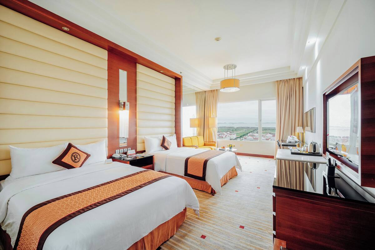 A hotel room with two beds, a desk, a chair, a TV, and a large window offering a city view.