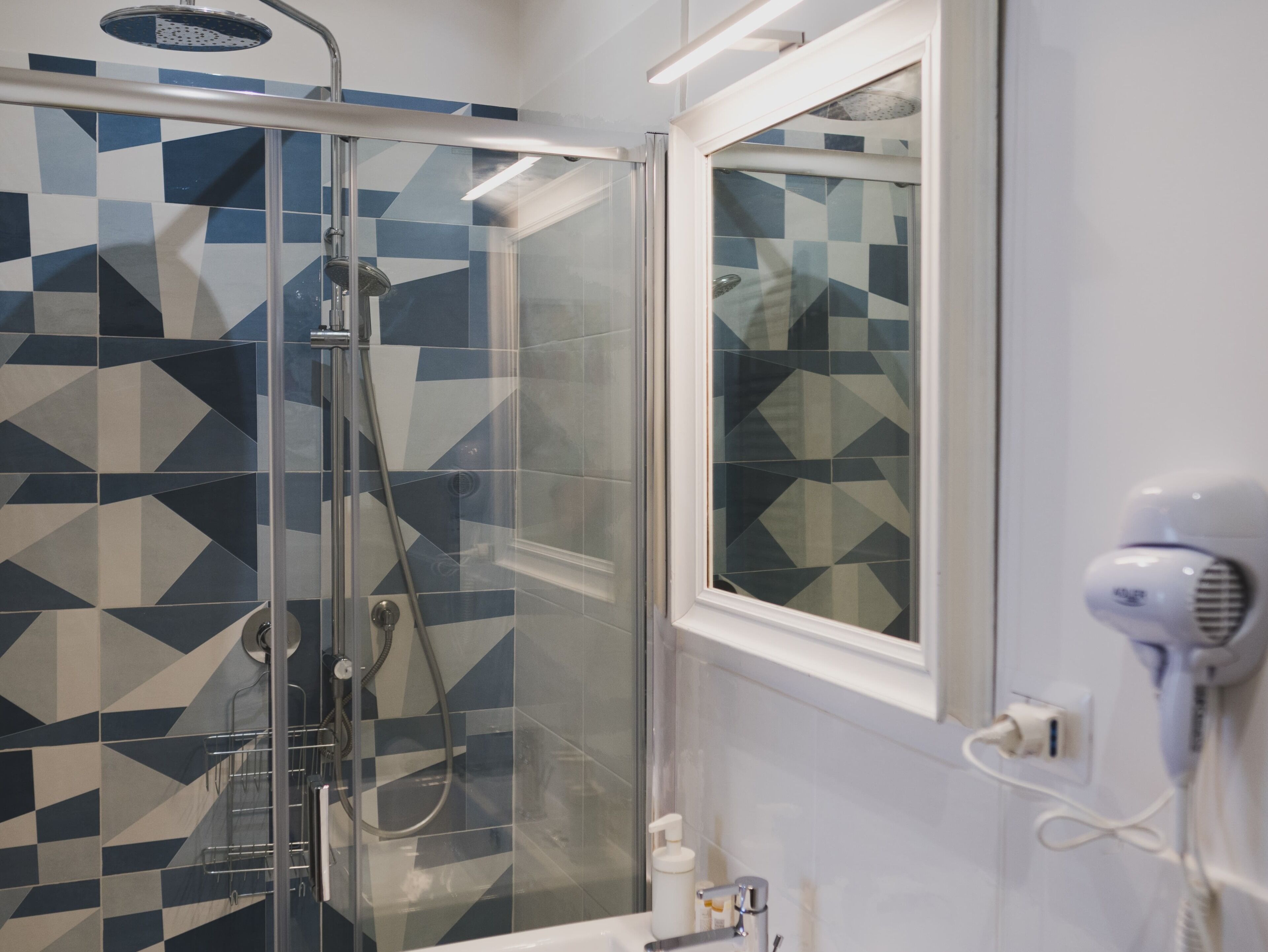 A modern bathroom with a glass-enclosed shower, geometric patterned tiles, a window, and a hairdryer on the counter.