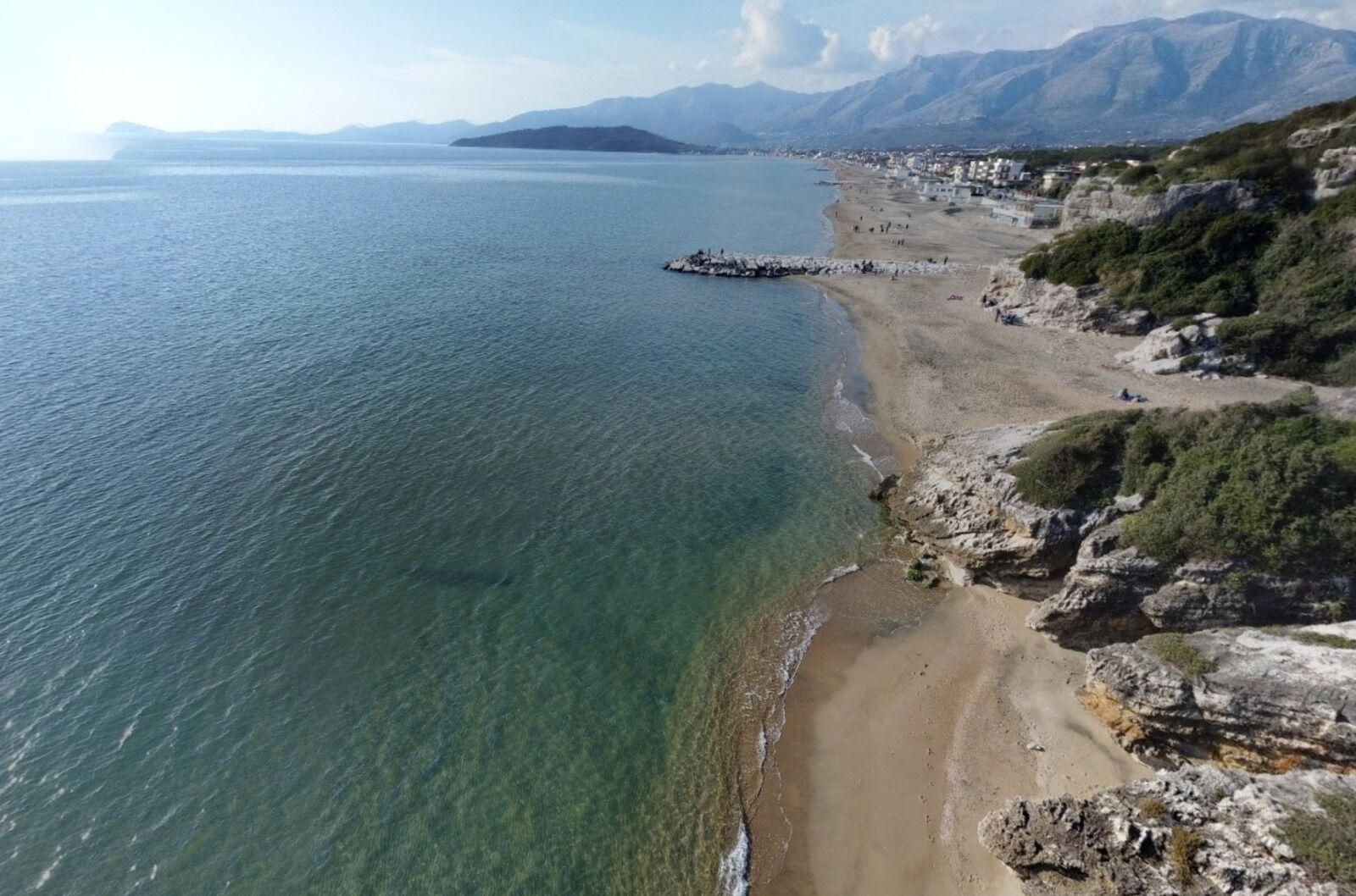 A coastal beach with clear waters, rocky cliffs, and a distant mountain range.