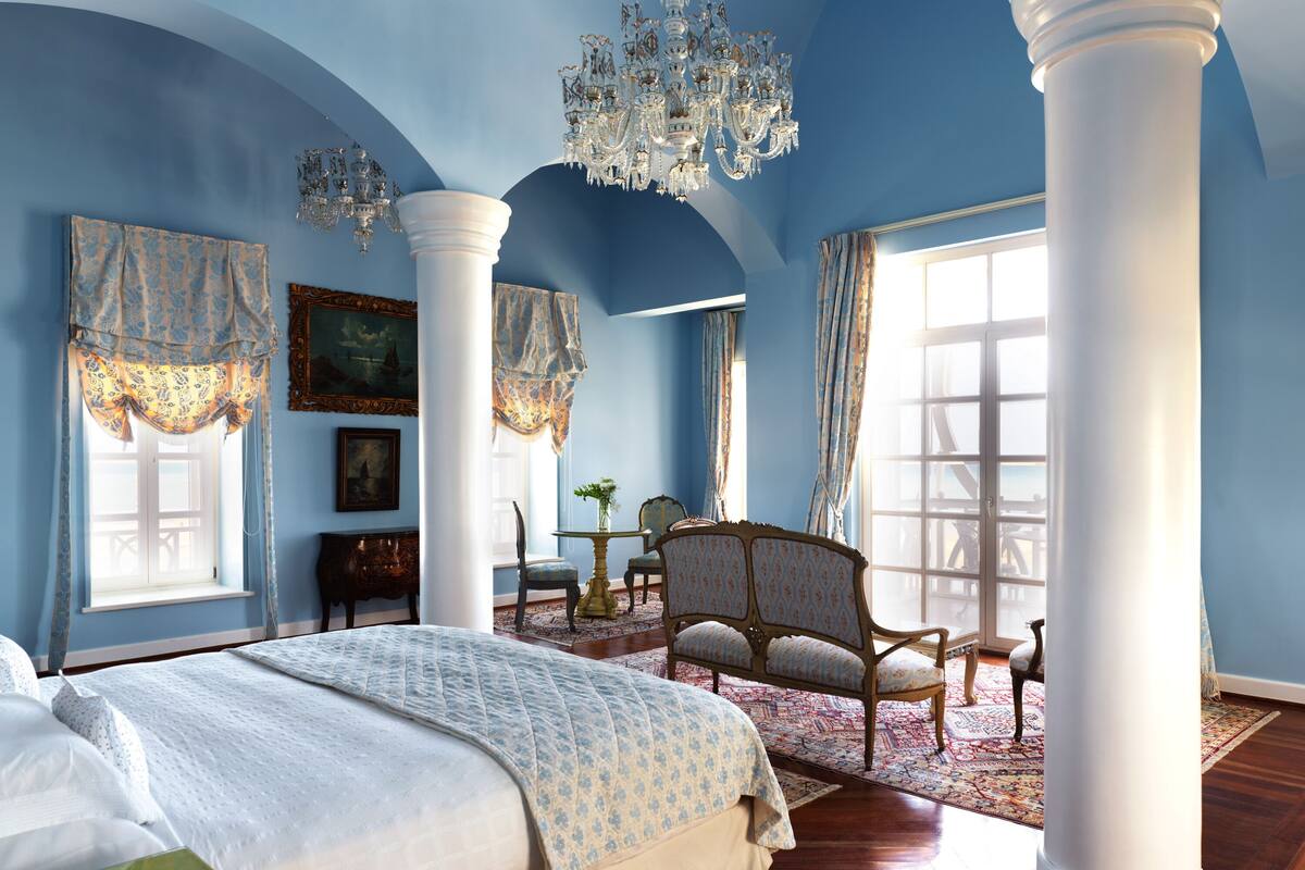 Un dormitorio con una cama grande, un candelabro, una silla y vistas al mar a través de una ventana.
