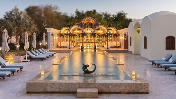 A pool area with lounge chairs, a decorative fountain, and a building with arches and lights. - La Maison Bleue (El Gouna)