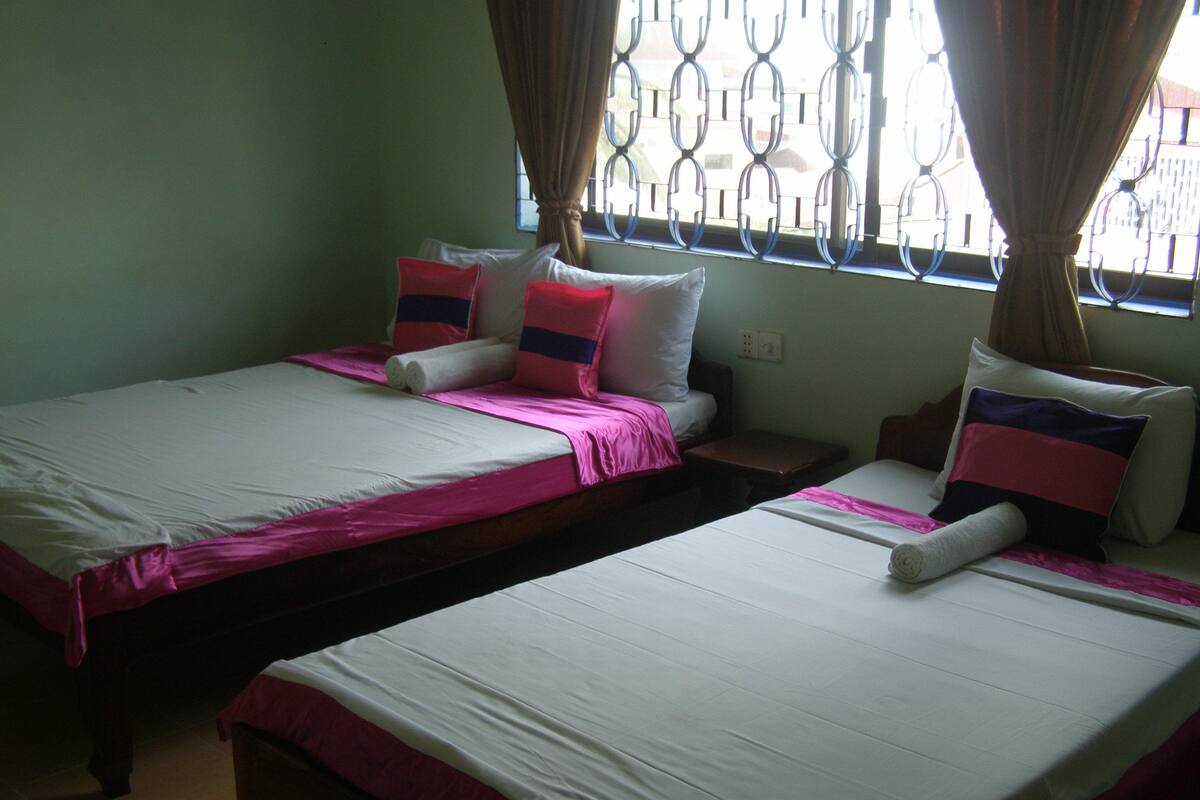 A room with two single beds, each with a pink bedspread and white pillows. There is a window with metal bars and curtains, and a small wooden nightstand between the beds.