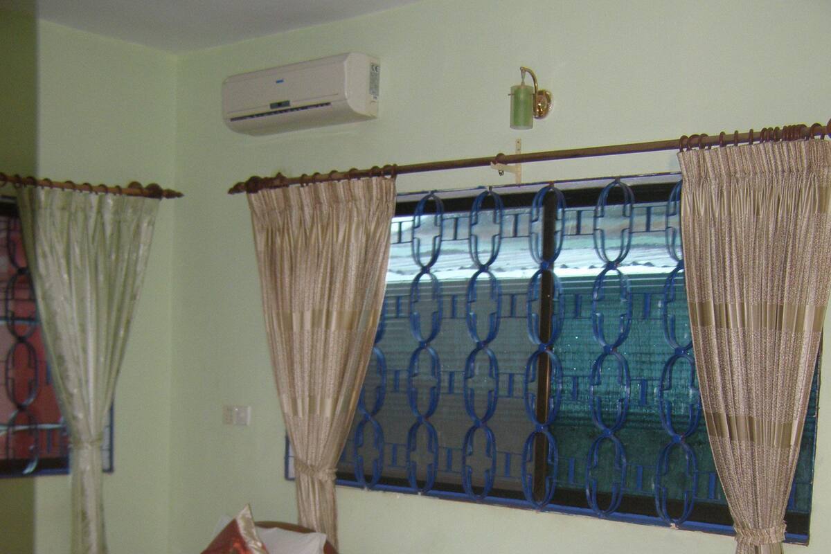 A room with two wooden beds, each with a pillow and a rolled-up towel. There is an air conditioning unit on the wall, and a window with blue metal bars and curtains.