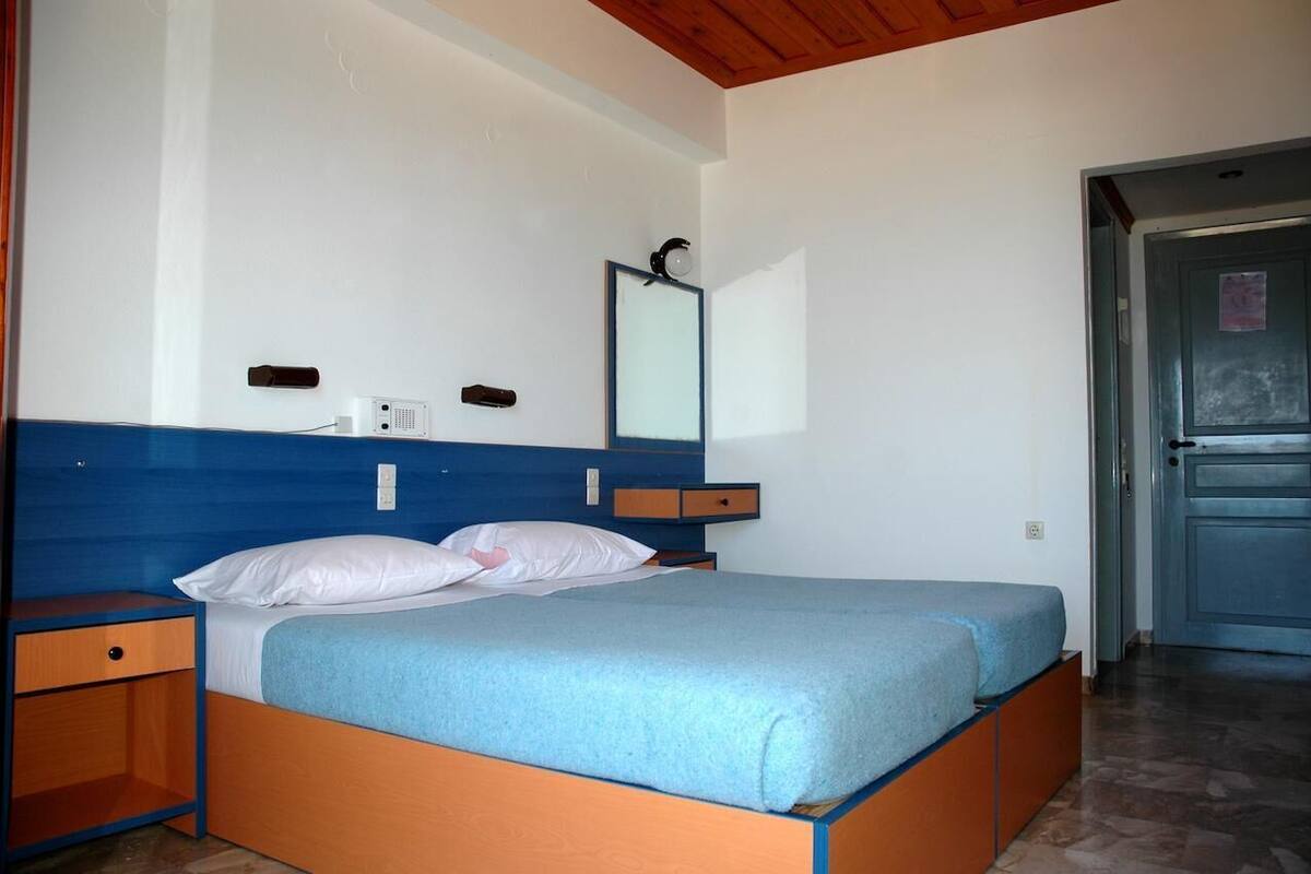 A single bed with a blue mattress and white pillows, a bedside table, a small drawer, a wall-mounted mirror, and a door to the right.