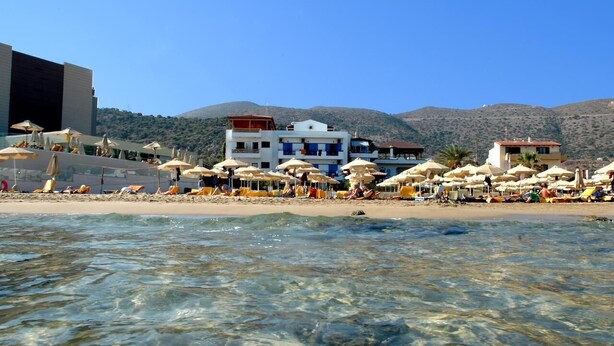 A beach with clear water, umbrellas, and buildings in the background. - Vlachakis Hotel (Hersonissos)