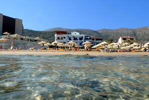 A beach with clear water, umbrellas, and buildings in the background. - Vlachakis Hotel (Hersonissos)