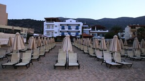 A beach with rows of sun loungers and umbrellas, a white building with balconies, and mountains in the background. - Vlachakis Hotel (Hersonissos)