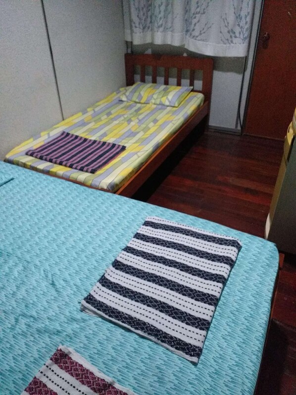 A small, compact room with two single beds, a wooden headboard, a window with curtains, and a wooden floor.