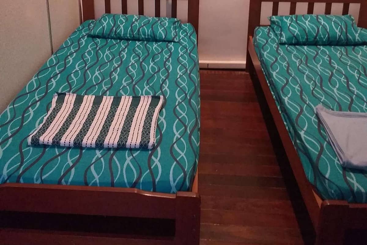 Una habitación con dos camas de madera, cada una con ropa de cama color turquesa estampada y un cojín a rayas. Se observa una obra de arte con formas geométricas y círculos sobre las camas.
