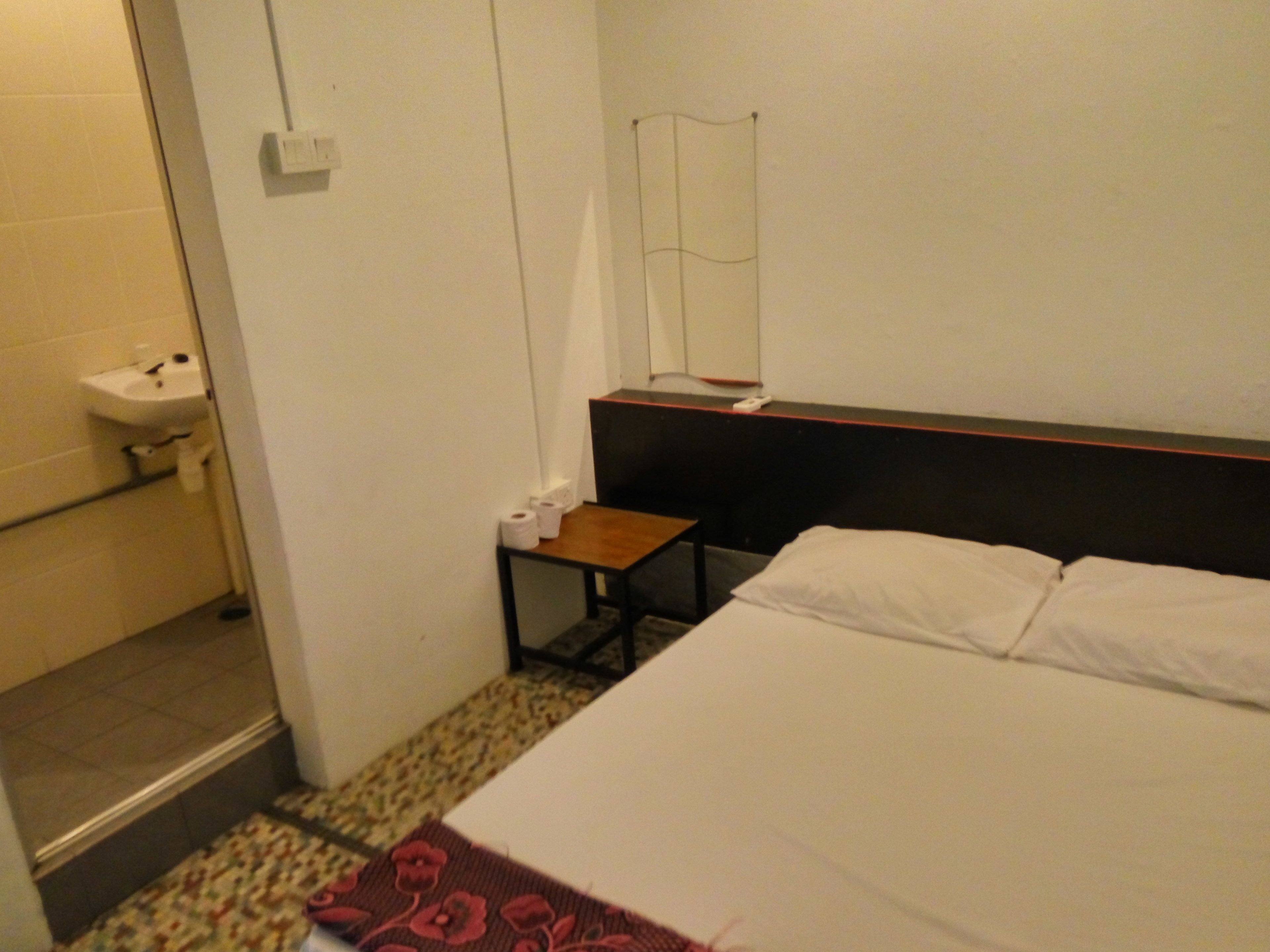 A hotel room with a bed, a small table with toiletries, a mirror, and a bathroom with a sink and toilet.