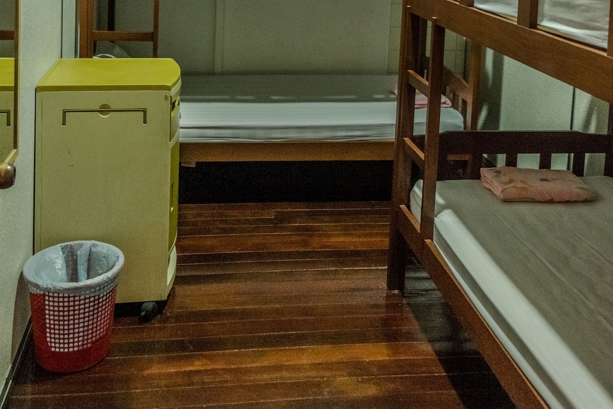 Dormitorio con literas, suelo de madera, un armario pequeño y un cubo de basura.