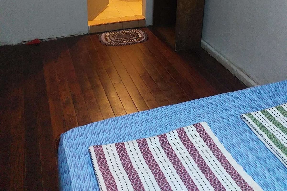 Una habitación pequeña y estrecha con piso de madera, una cama azul con cubrecama a rayas, un lavamanos blanco y un estante de madera oscura.