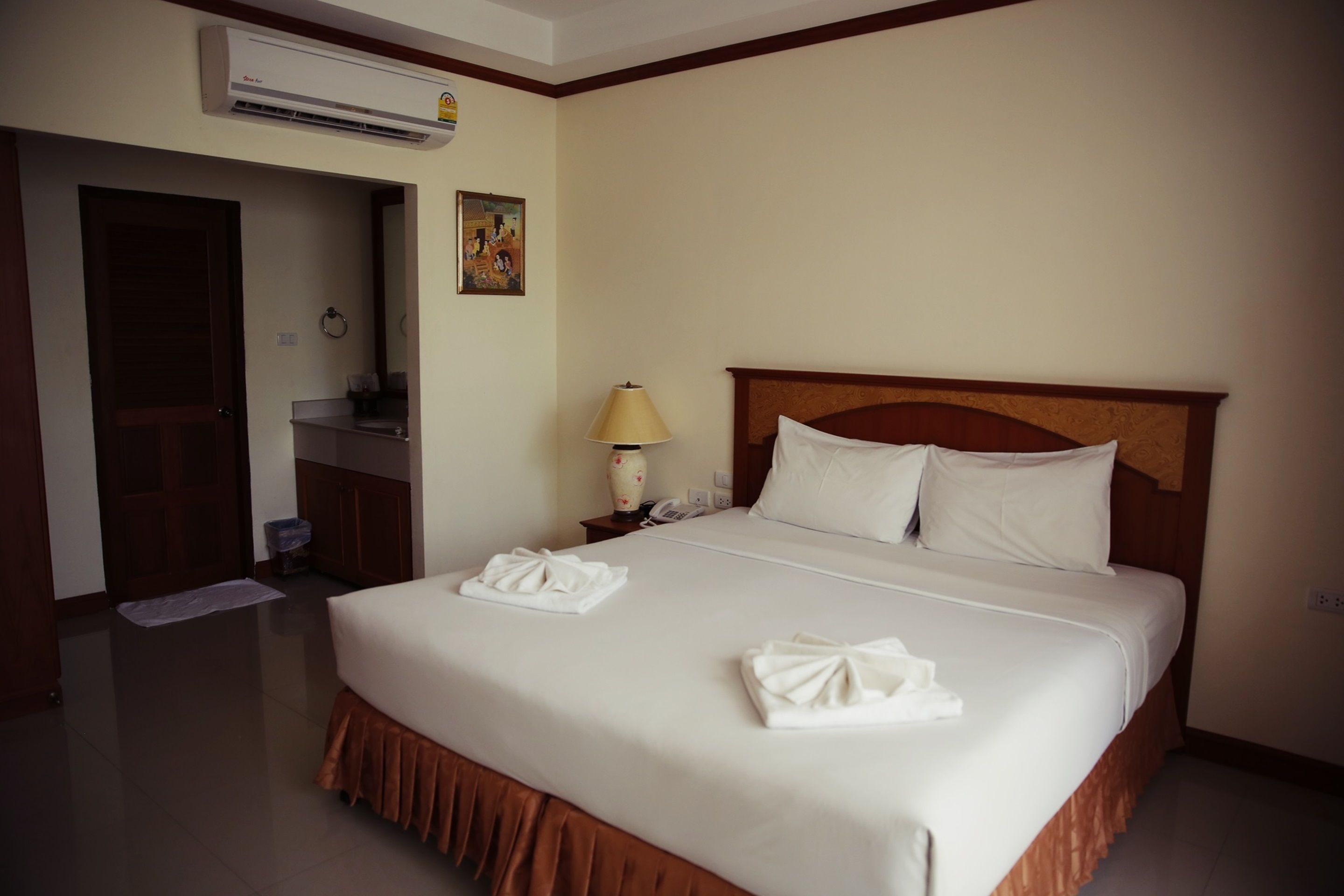 A hotel room with a large bed, a wooden headboard, a nightstand with a lamp, and an air conditioning unit on the wall.