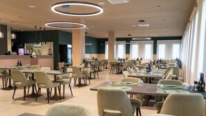 Un restaurante amplio con numerosas mesas y sillas, una barra con un empleado, y grandes ventanales que permiten la entrada de luz natural.