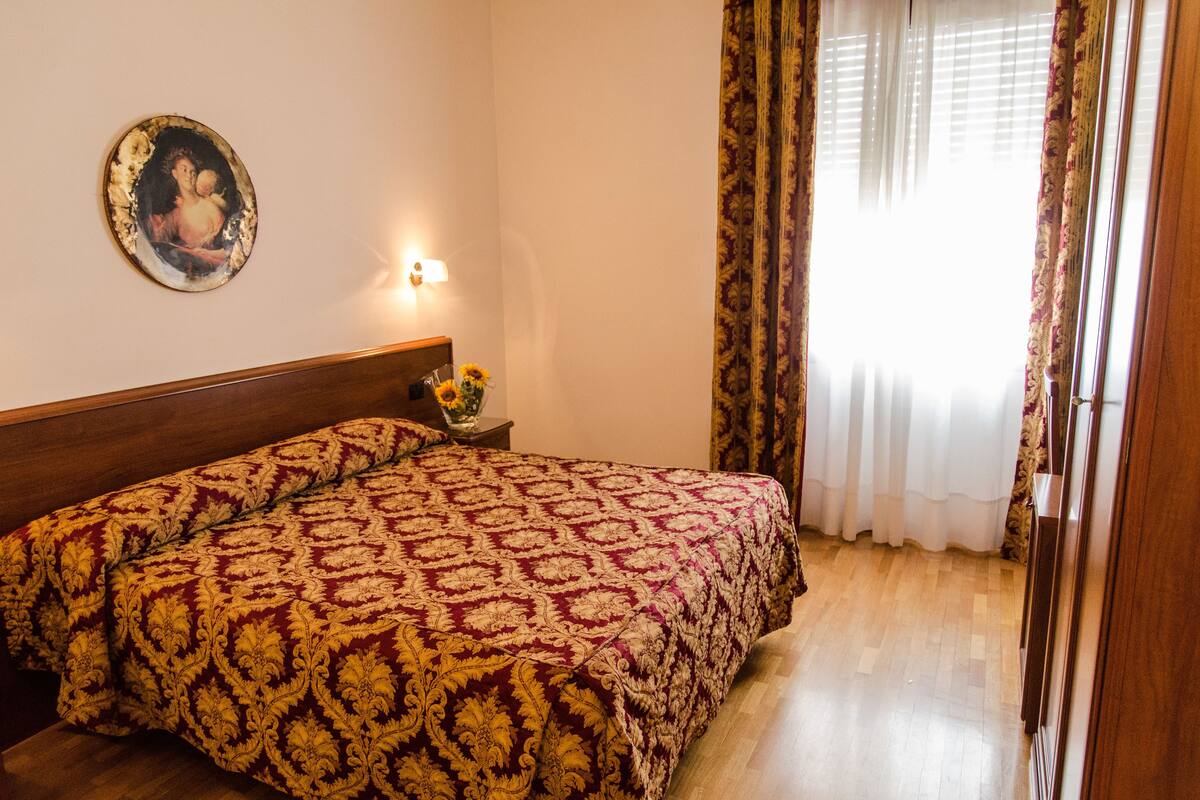 A bedroom with a wooden headboard, a patterned bedspread, a nightstand with a vase of sunflowers, and a painting on the wall.